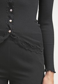 Czarny prążkowany kardigan z falbaniastą koronkową lamówką, pięcioma guzikami i dopasowanymi długimi rękawami. Zestawiony z czarnymi wąskimi spodniami.