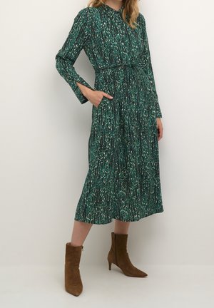Kobieta w zielonej, wzorzystej sukience midi z długimi rękawami i talią zwężoną, w połączeniu z brązowymi zamszowymi botkami na obcasie.