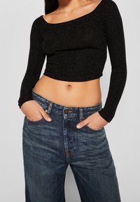 Musta pitkähihaine crop-top, jossa on hienovarainen kimmellys, yhdistetty löysiin sinisiin farkkuihin, joissa on haalistunut kuvio ja kulutettuja yksityiskohtia.