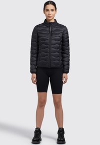 Černá vycpaná bunda s vysokým límcem, která má texturovaný vlnitý design a přední zip. Nosená přes černé proužkované šortky a tenisky.