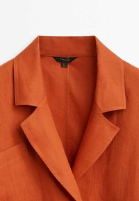 Brązowy lniany blazer w kolorze rdzy z klapami, pojedynczą kieszenią na piersi i minimalistycznym wzornictwem. Metka wskazuje rozmiary S, M, L oraz instrukcje dotyczące pielęgnacji.