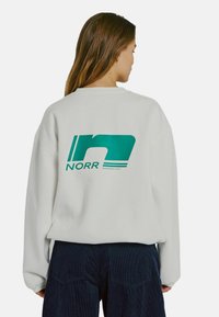 Biely oversized sveter s zelenou grafickou potlačou "NORR" na chrbte. Textúrovaná tkanina s rebrovanými manžetami a voľným strihom.