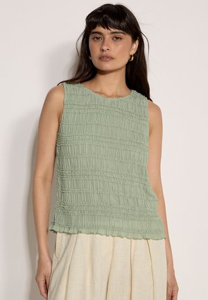 Joven mujer con el cabello largo y oscuro, lleva pendientes de aro dorados, una blusa sin mangas de color verde claro con textura y pantalones beige de talle alto.