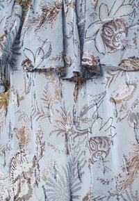 Tejido de gasa azul claro con patrones florales en tonos de marrón, dorado y plata. El diseño incluye detalles con volantes y una textura suave.