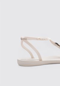 Sandalia de plástico blanca con suela plana, que presenta un diseño de tira en T y hardware decorativo en el tobillo. Textura suave con acentos mínimos.