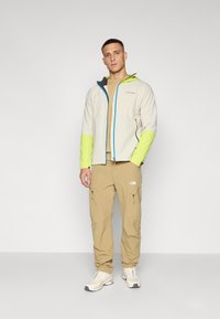 Jachetă impermeabilă gri deschis cu mâneci verzi aprinse și fermoar albastru contrastant, purtată cu pantaloni cargo bej și adidași bej.