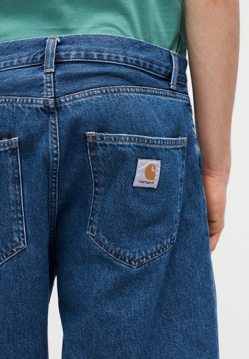 Vista trasera de una persona vistiendo jeans Carhartt de mezclilla azul con bolsillos traseros visibles y parche con el logo de Carhartt en el bolsillo derecho.