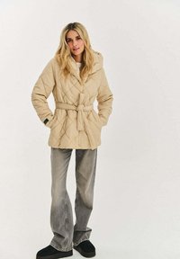 Béžová prošívaná bunda s kapucí, páskem v pase a bočními kapsami. Kombinováno s šedými rozšířenými džíny a černými botami na platformě.