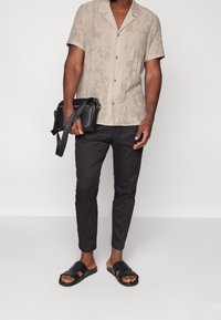 Beżowa koszula z krótkim rękawem w liściasty wzór, czarne spodnie i czarne sandały. Trzyma czarną torbę na ramię.