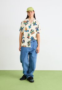 Cămașă crem cu mânecă scurtă, decorată cu modele colorate, asociată cu blugi lejeri de culoare albastră deschisă și adidași negri. Se poartă o șapcă verde și un colier din mărgele.