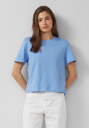 Femme aux longs cheveux bruns portant une chemise bleu clair à manches courtes et un pantalon blanc, debout contre un mur gris uni.