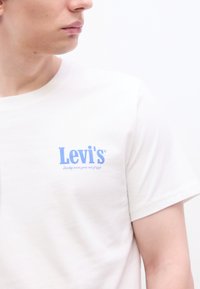 Νέος άνθρωπος που φοράει λευκό κοντομάνικο Levi's με μπλε λογότυπο και σύνθημα, κοιτώντας προς τα αριστερά τους μπροστά σε απλό λευκό φόντο.