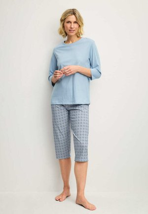 Blonde vrouw staat blootsvoets tegen de muur, draagt een lichtblauw shirt met driekwart mouwen en een broek met patroon tot aan de knie.