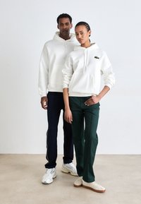 Fehér kapucnis pulóverek kenguruzsebekkel és zöld Lacoste logóval, sötétkék és zöld nadrágokkal párosítva. A modellek világos betonpadlón állnak.