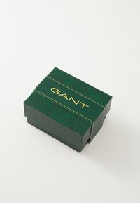 Zelená obdĺžniková krabička s reliéfnym paisley vzorom, zlaté logo "GANT" na vrchu, bezpečné uzavretie, vyrobená z textúrovaného kartónu.