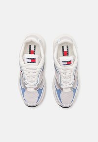 Pár bílých a světle modrých tenisek Tommy Jeans s síťovinou a logovými nášivkami na jazycích a bocích, viděno zeshora.