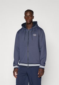 Fato de capuz navy com fecho em zip, capuz com cordão, punhos e bainha em detalhes brancos. Apresenta o logótipo EAZ no peito. Textura suave.