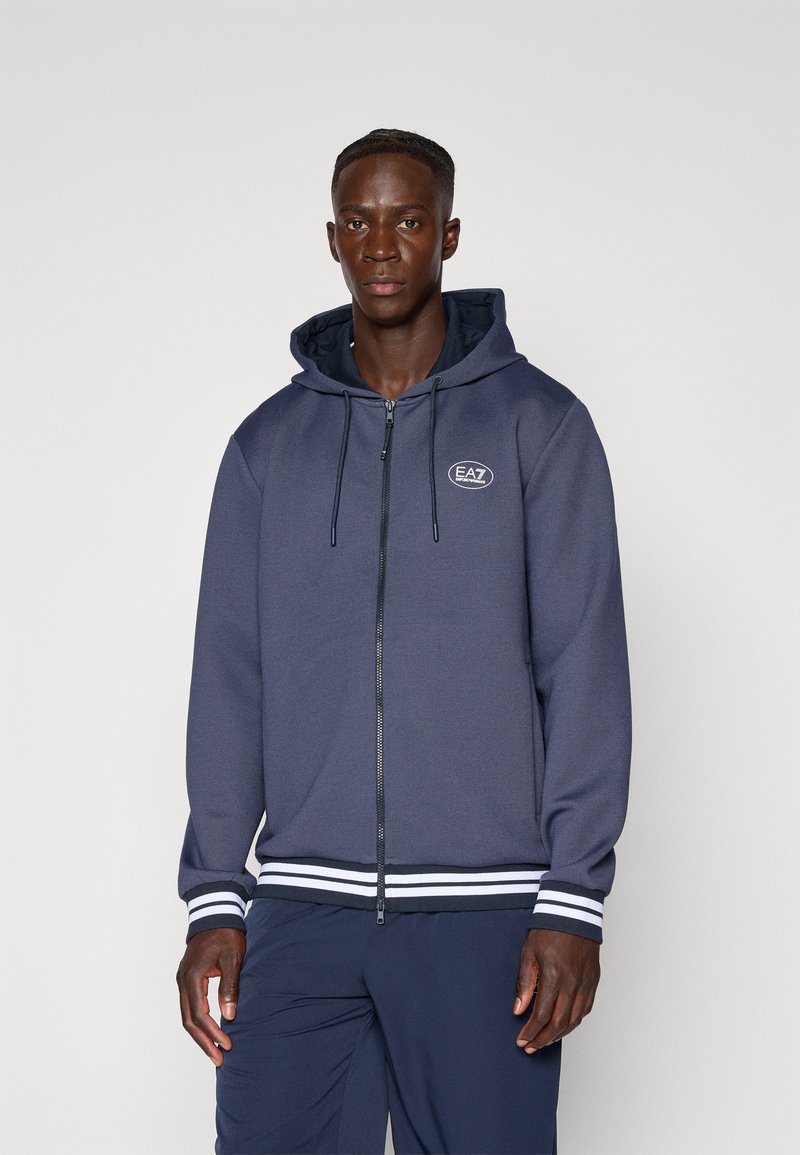 Fato de capuz navy com fecho em zip, capuz com cordão, punhos e bainha em detalhes brancos. Apresenta o logótipo EAZ no peito. Textura suave.