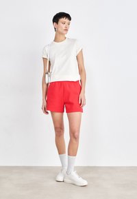 T-shirt bianca a maniche corte con dettagli del logo, abbinata a pantaloni corti rossi. Il modello indossa scarpe da ginnastica bianche e calze al ginocchio. Design semplice e casual.