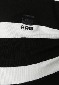 Schwarz-weißer gestreifter Stoff mit gerippter Textur, versehen mit einem kleinen, dunklen Logo-Patch mit dem Wort "RAW". Schlichter Ausschnitt.