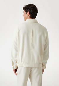 Light-colored, smooth fabric jacket with a high collar, elastic waistband, and a back vent detail. Long sleeves and minimal hardware accents.