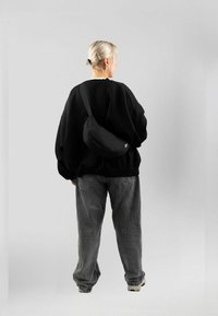 Borsa a tracolla nera realizzata in materiale morbido, indossata a tracolla su una felpa nera dalla vestibilità ampia e jeans grigi larghi. Sfondo neutro.