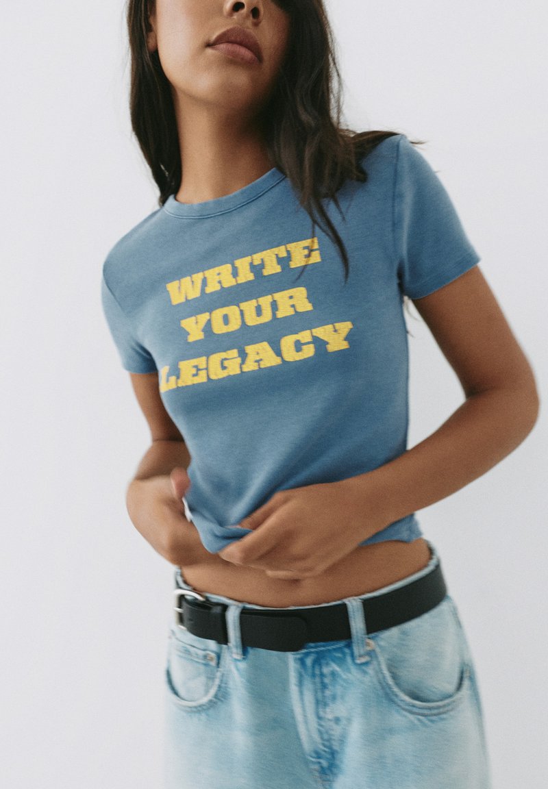 Woman with long dark hair wearing a blue t-shirt with yellow text "WRITE YOUR LEGACY" and light blue jeans with a black belt.