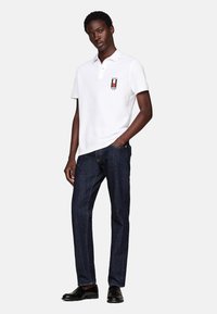 Polo blanca con una tapeta de dos botones y un acento de logo bordado. Combinada con jeans de mezclilla oscura y zapatos negros.