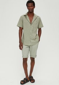 Kurzärmliges, hellgrünes Hemd mit kleinen, gemusterten Akzenten, kombiniert mit hellgrünen Shorts und schwarzen Rutschsandalen. Schlichter, lässiger Look.
