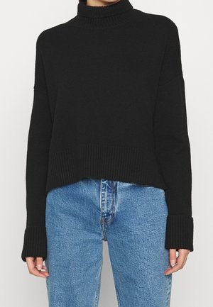 Person iført en sort ribbet turtleneck sweater med lange ærmer og manchetter, parret med højhævede blå jeans.