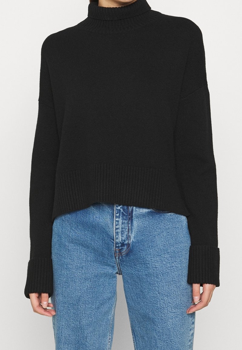 Persona che indossa un maglione a collo alto a coste nere con maniche lunghe e dettagli sui polsini, abbinato a jeans blu a vita alta.