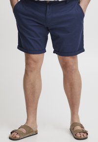 Mørkeblå bomullsshorts med rullet kant, sidelommer og knappelukking. Kombinert med brune semskede sandaler med justerbare stropper.