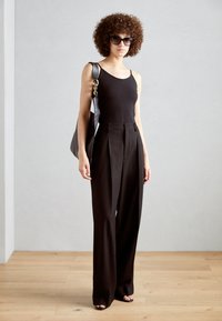 Black tank top, high-waisted, pleated black trousers, oversized black sunglasses, and a large black tote bag. Wood floor, minimal background.