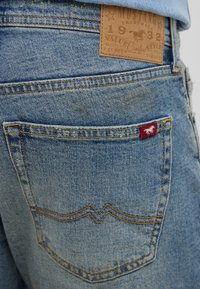 Jasnoniebieskie dżinsy z tyłu mają kieszeń z czerwoną metką logo. Materiał ma zużytą teksturę, a widoczne szwy podkreślają detale.