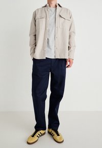 Beżowa koszula na guziki z dwoma przednimi kieszeniami, pod nią szara koszulka, granatowe spodnie i żółte trampki z czarnymi paskami.