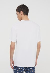 T-shirt bianca a maniche corte con colletto tondo, realizzata in cotone, con una texture liscia. Una persona la indossa, vista da dietro.