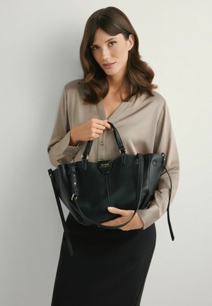 Kobieta o falowanych brązowych włosach ubrana w beżową bluzkę i czarną spódnicę, trzyma czarną skórzaną torbę na ramię z krótkimi uchwytami i paskiem na ramię.