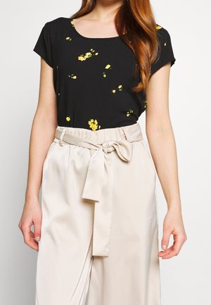 Femme portant un haut noir à manches courtes avec un petit imprimé floral jaune et un pantalon crème taille haute noué avec une ceinture en tissu.