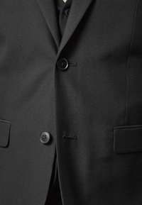 Svart blazer med ensidig knäppning, två svarta knappar och sidofickor. Slät tyg med subtil textur och kontrasterande sömmar.