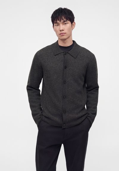 Jeune homme aux cheveux courts noirs portant un cardigan gris foncé boutonné et un pantalon noir, debout les mains dans les poches sur fond blanc.