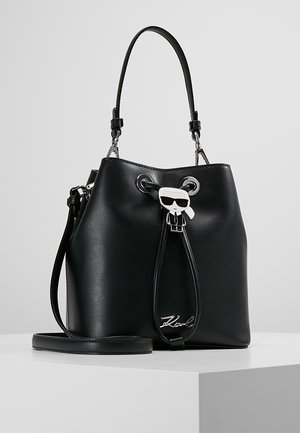 Schwarze Lederbeuteltasche mit Tragegriff oben, verstellbarem Schulterriemen und einem Anhänger mit einer stilisierten Figur, die eine Sonnenbrille trägt, sowie einem charakteristischen Detail.