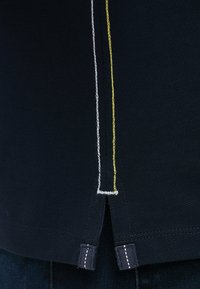 Navy polo shirt section with textured fabric. Features white and yellow stitching along the seams and side slits at the hem.