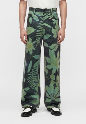 Grønne og sorte bukser med vide ben og stort løvformet botanisk print, båret med sorte og hvide loafers og en hvid skjorte.