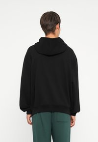 Persona de espaldas vistiendo una sudadera negra de gran tamaño con la capucha bajada y pantalones verdes holgados, frente a un fondo blanco liso.