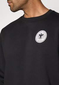 Melnā kokvilnas džemperis ar apaļu kakla izgriezumu un baltu apaļu logotipu ar uzrakstu "Deutscher Fussball Bund" un ērgļa dizainu.