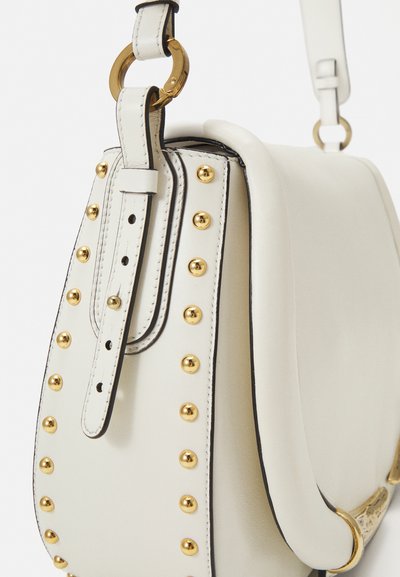 White leather handbag features gold studs along the side, a curved flap, and a gold metal ring on the strap. Smooth texture, structured design.