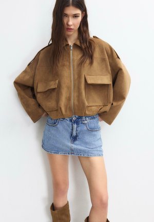 Junge Frau trägt eine braune, übergroße Jacke mit Reißverschluss und großen Taschen, einen blauen Jeansminirock und braune Stiefel, steht vor einer weißen Wand.