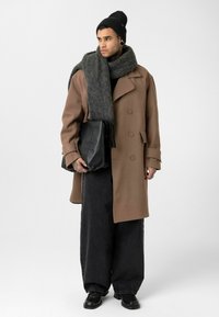 Man die een grote bruine jas draagt, zwarte wijde broek, zwarte muts, grijze sjaal, een grote zwarte leren tas vasthoudt, staand tegen een witte achtergrond.