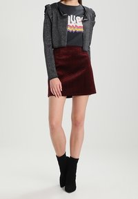 Cardigan corto scintillante argento su una t-shirt nera con grafica, abbinato a una gonna bordeaux con motivo a zigzag e stivaletti neri.