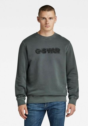 Junger Mann mit kurzen Haaren, der einen dunkelgrauen G-Star-Pullover und blaue Jeans trägt, vor einem weißen Hintergrund steht.